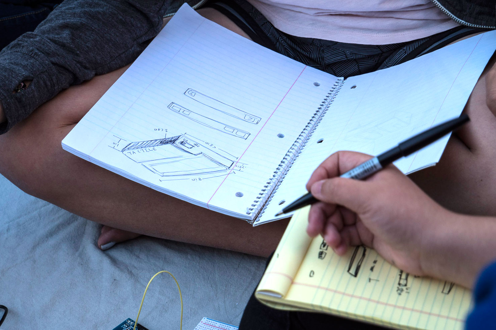 Photo of sketches of the Tactile device in notebooks that are resting on the laps of two students, one of them holding a pen